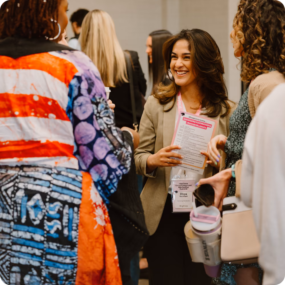 A woman with long dark hair, holding a booklet, smiles while engaged in conversation with two other women. They appear to be in a networking setting, surrounded by a lively group of people.