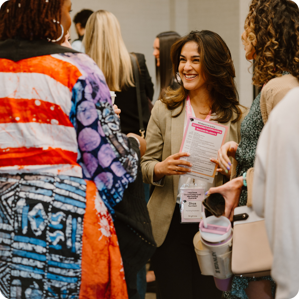 A woman with long dark hair, holding a booklet, smiles while engaged in conversation with two other women. They appear to be in a networking setting, surrounded by a lively group of people.