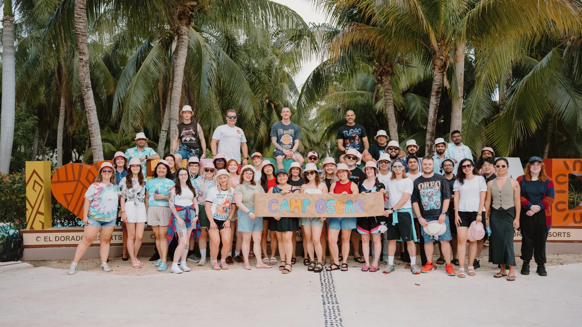 A large group of people is gathered outdoors in front of palm trees, holding a banner that says 'CAMP OSCAR.' They appear to be on a casual outing, wearing relaxed clothing.