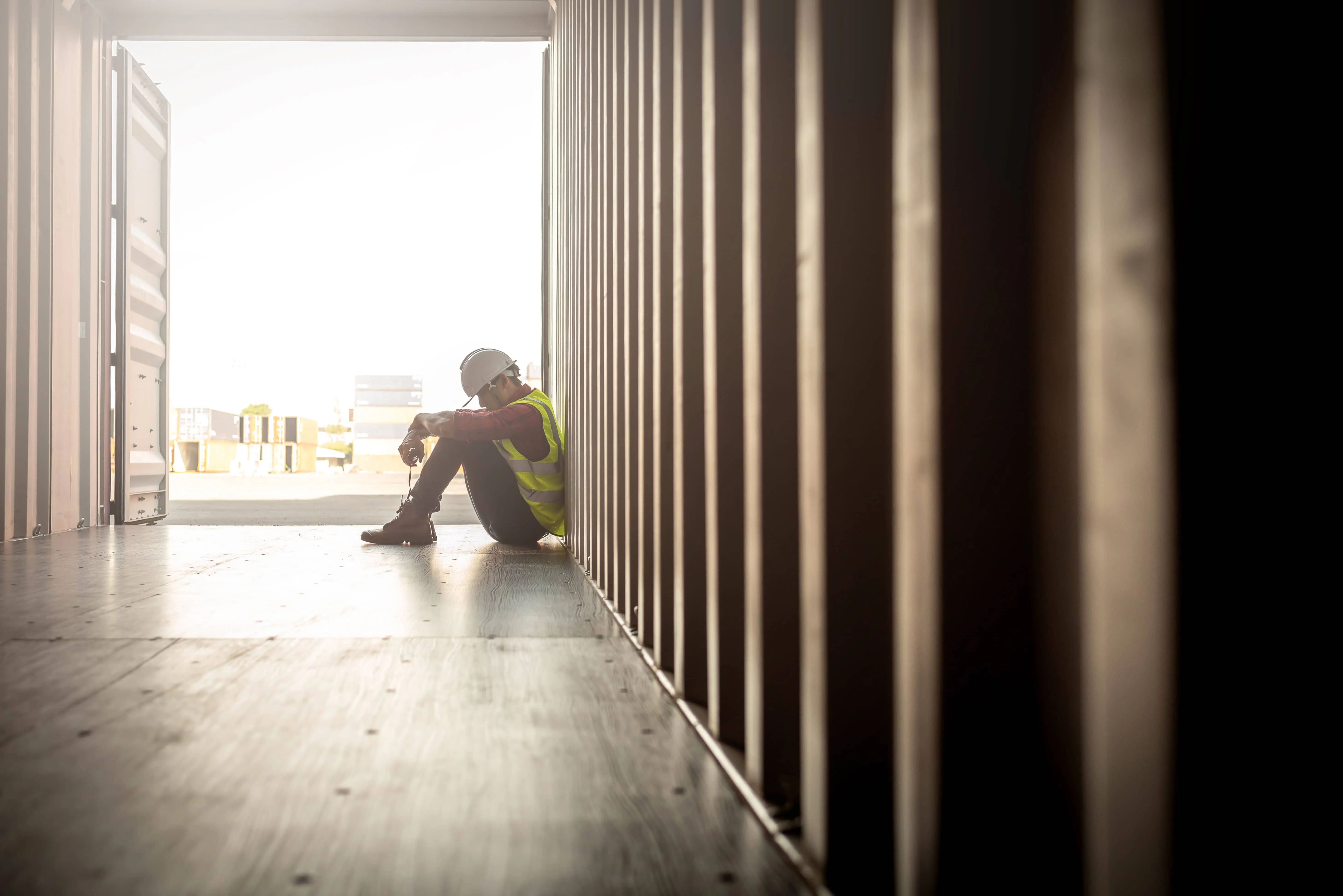 Construction worker in safety vest and helmet sitting on the floor inside a shipping container, leaning against the wall with head down.