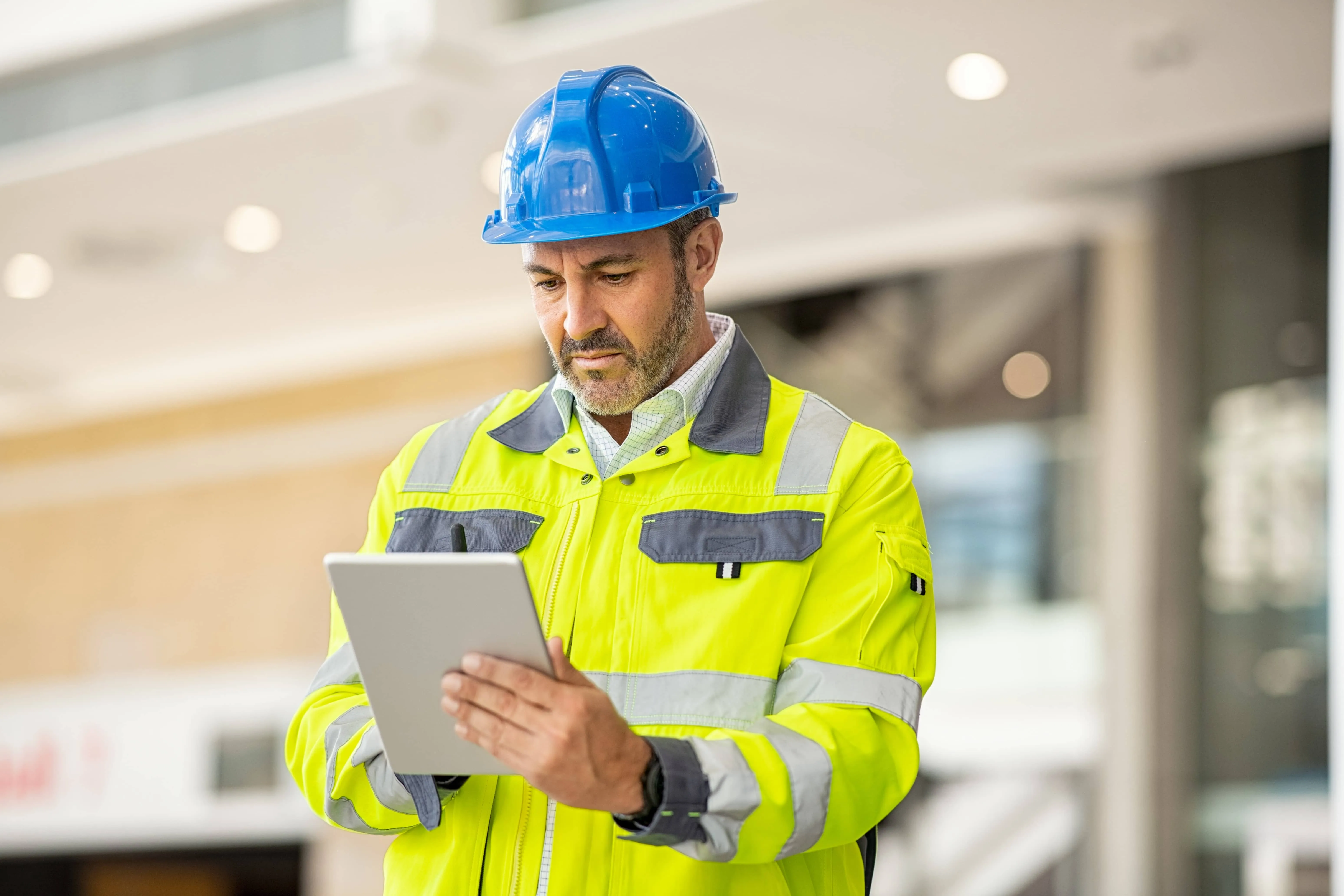 Male construction worker in a blue hard hat and yellow reflective jacket using a digital tablet indoors.