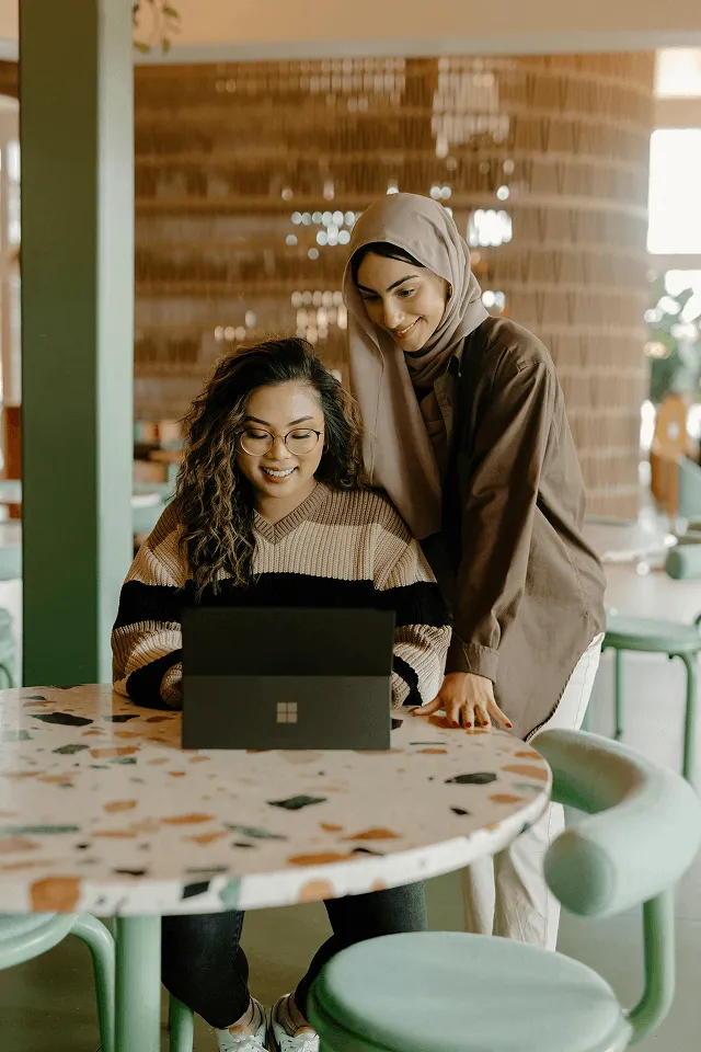 Two women collaborating at a table with a laptop, one sitting and the other standing and smiling.