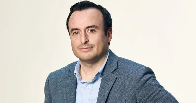 Man wearing a light blue shirt and gray blazer against a plain light background.