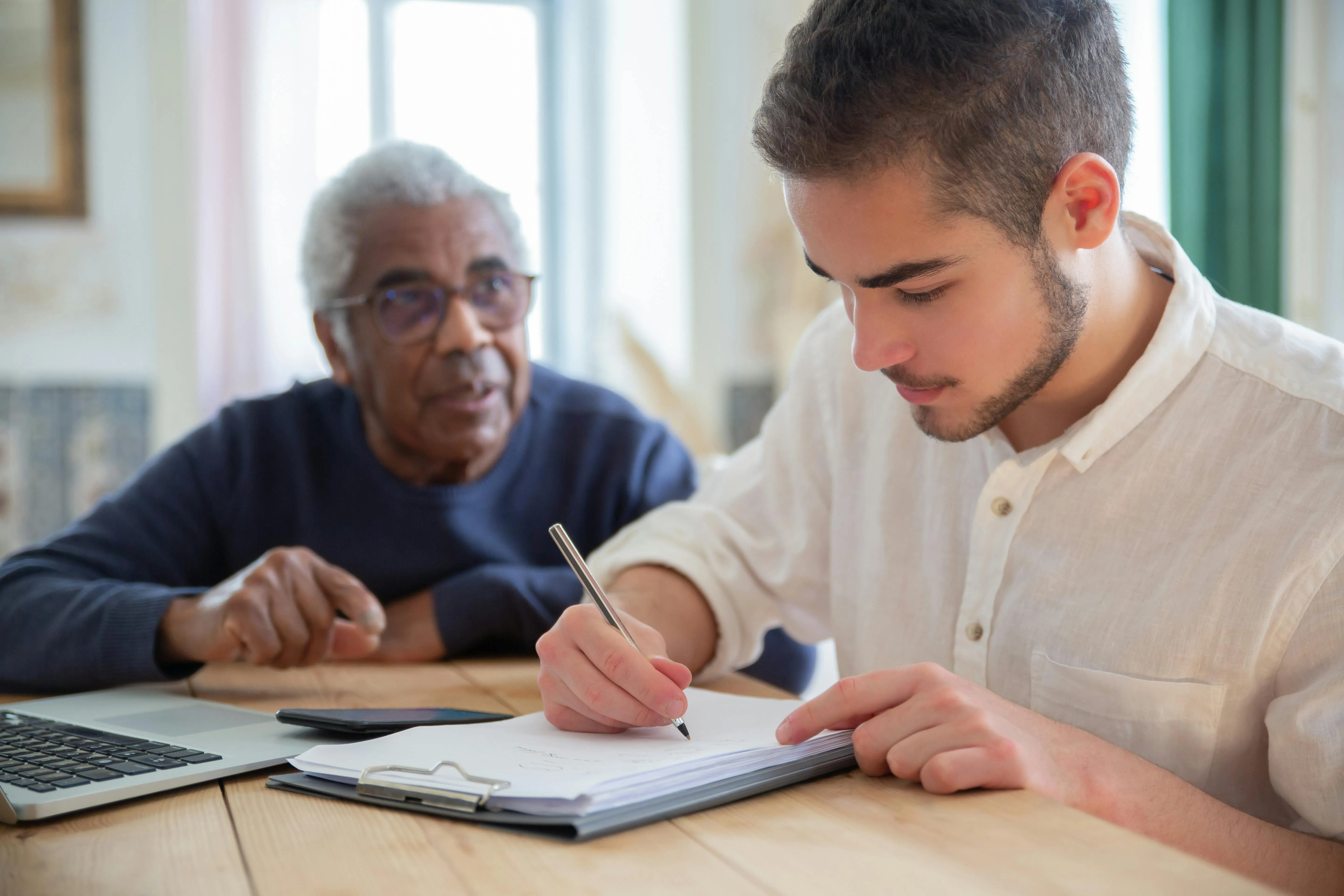 Young man in white shirt writing on a clipboard while an elderly man in glasses watches attentively.
