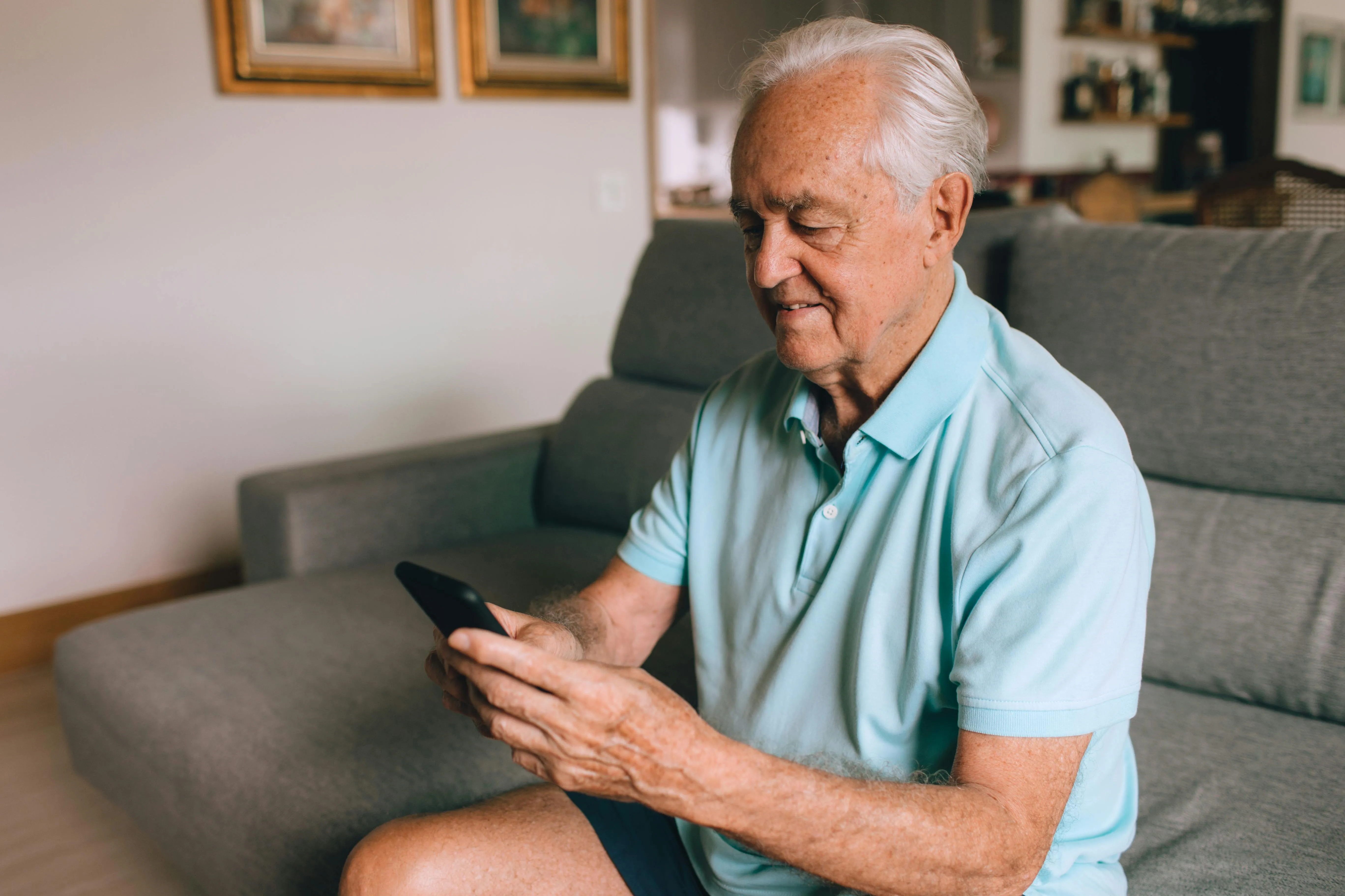 Elderly man sitting on a gray couch, looking at and holding a smartphone with a slight smile.
