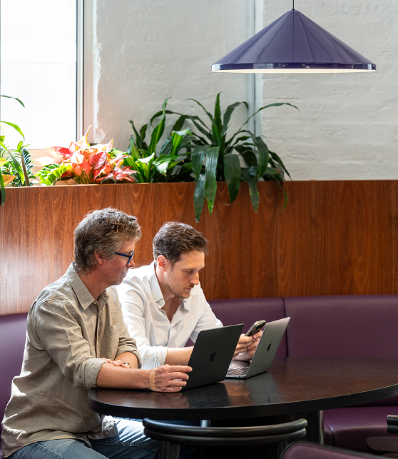 Two Q-CTRL colleagues at a round table in a modern office lounge, each using a laptop with one checking his phone, surrounded by plants and a purple hanging lamp.