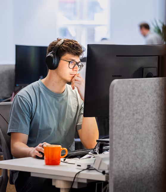 Young man wearing glasses and headphones working intently at a computer in an office space with a bright window in the background.