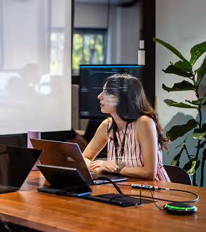Woman in striped sleeveless top sitting at a desk with a laptop in front of a computer monitor in a modern office.