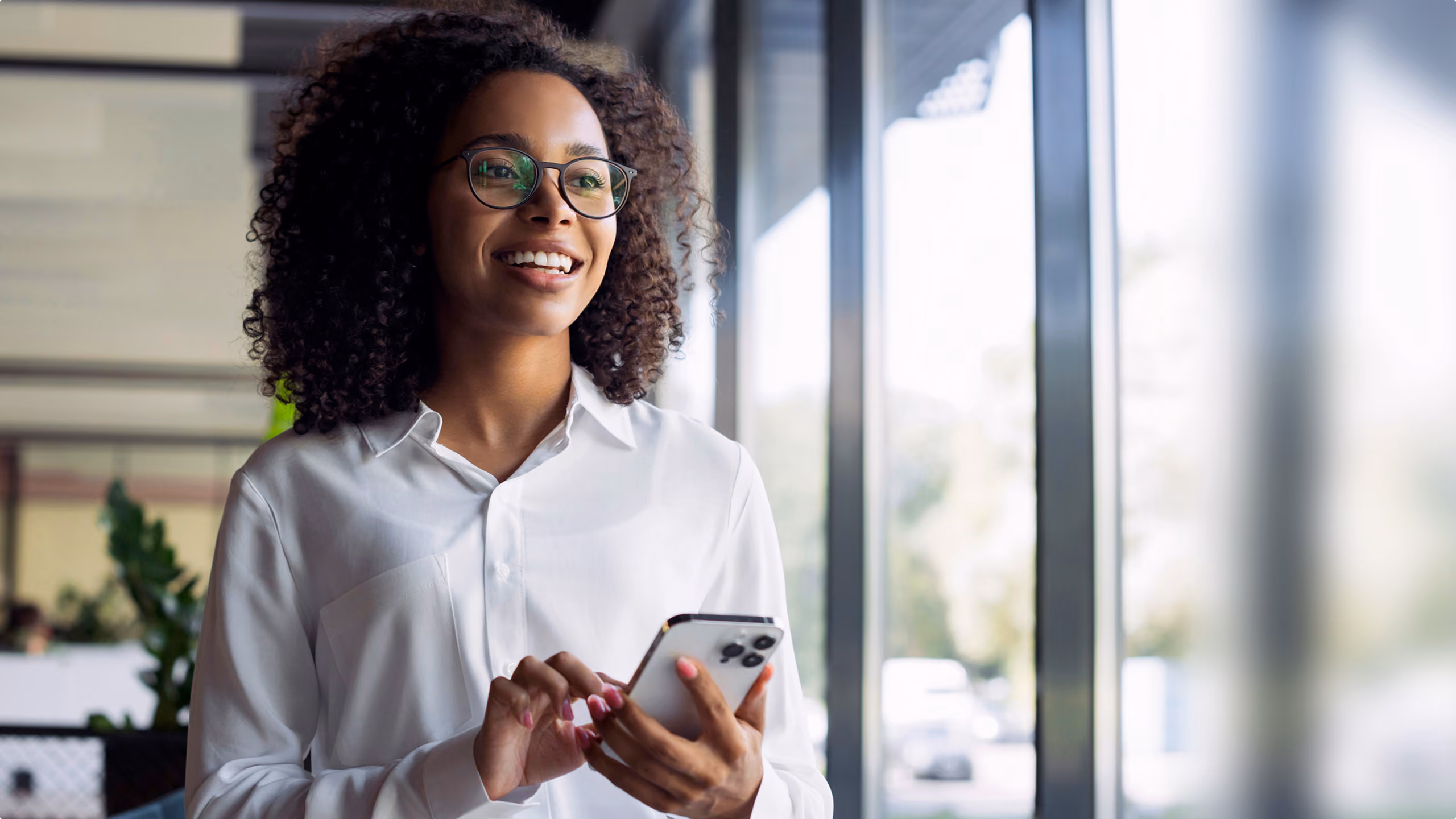 Smiling woman with curly hair and glasses using a smartphone near large windows in an office.