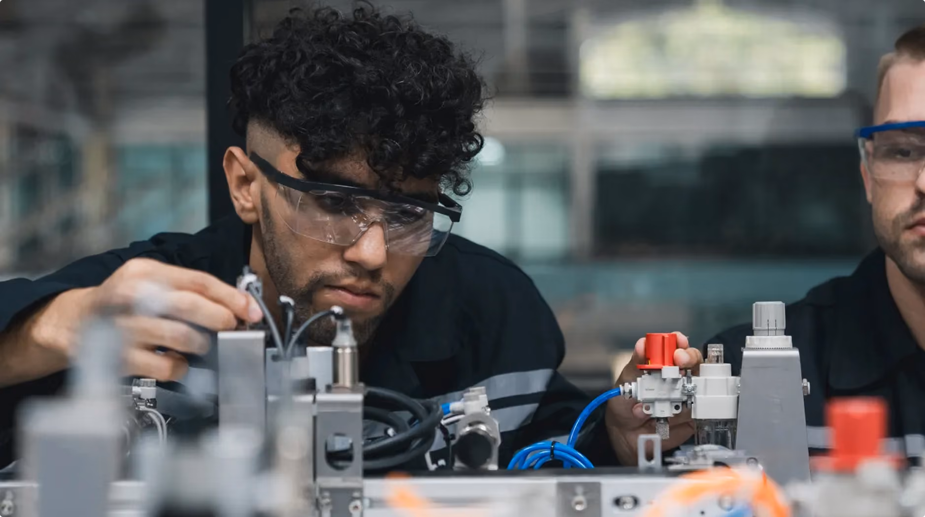 Two engineers wearing safety glasses adjusting machinery with wires and valves in a technical workspace.