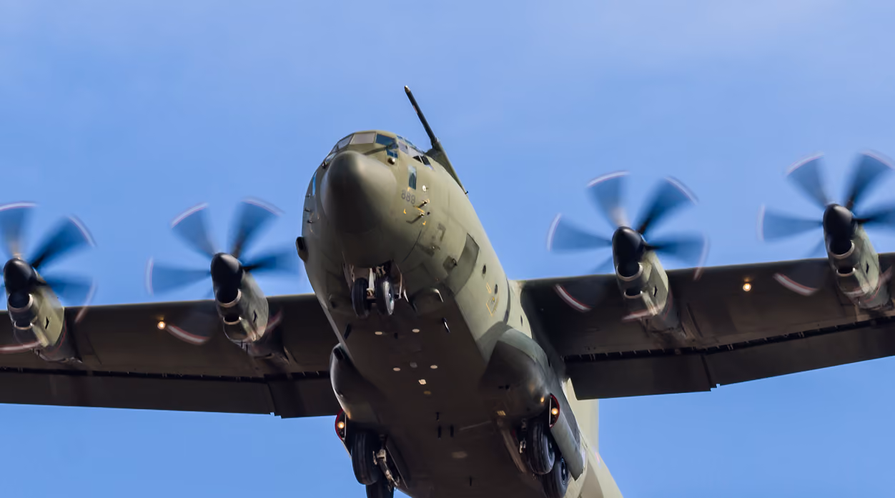 Military transport plane with four spinning propellers flying low against a clear blue sky.