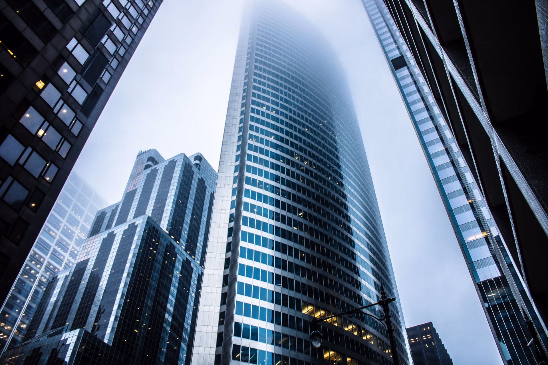 Skyscrapers with glass facades in foggy weather viewed from street level looking up.