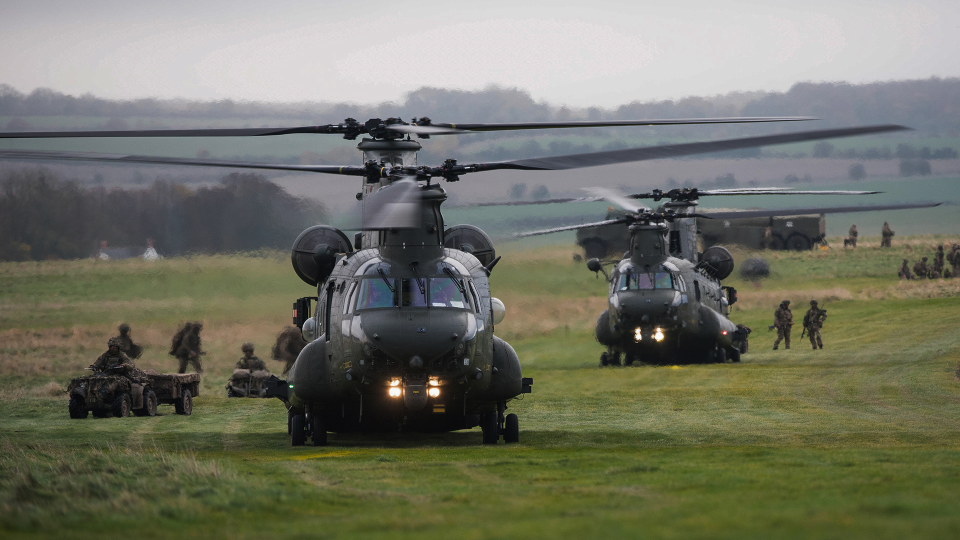 Two military helicopters on grass field with soldiers and vehicles surrounding them.