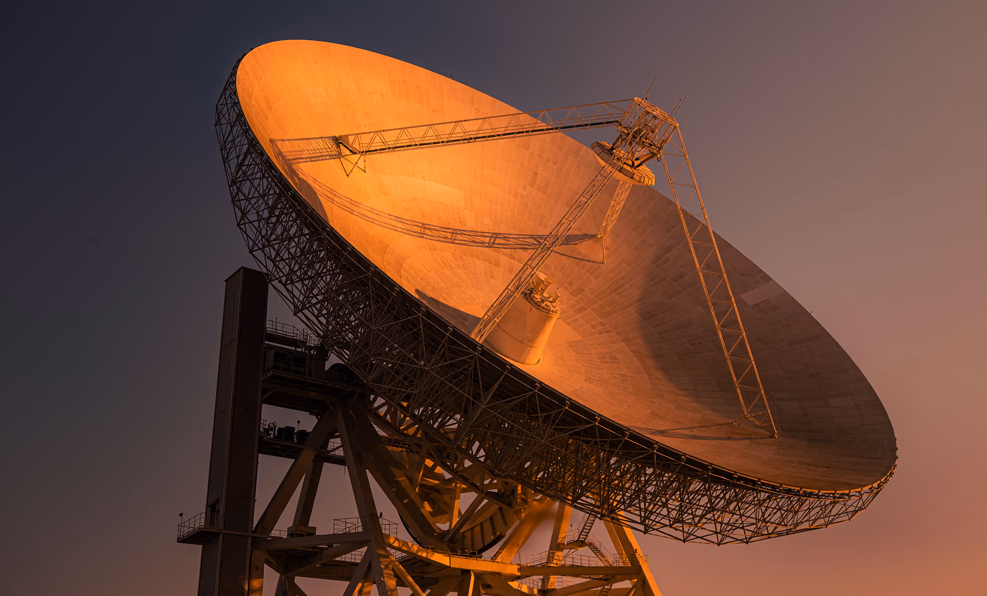 Large radio telescope dish bathed in orange light against a dusk sky.