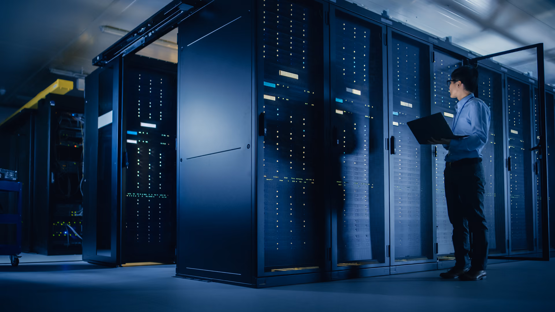 Man with laptop standing in a data center next to server racks with blinking lights.