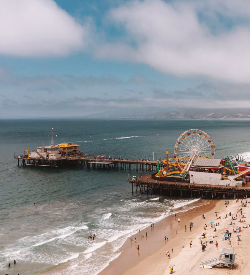 Crowded Santa Monica beach beside a pier with a Ferris wheel and amusement rides under a cloudy blue sky.