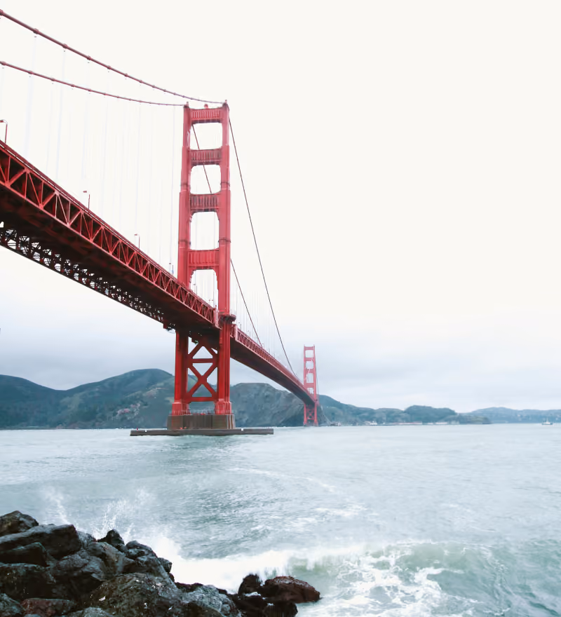 Red suspension Golden Gate Bridge over water with hills in the background under cloudy sky.