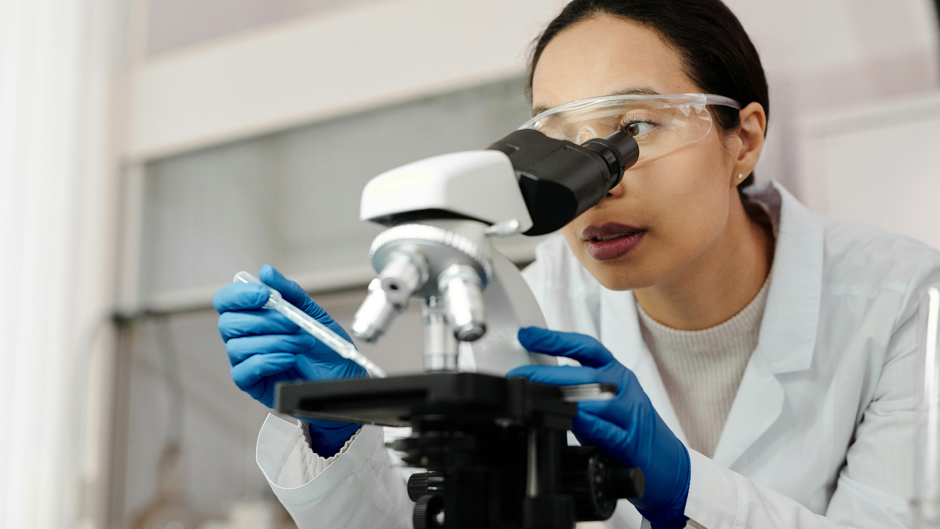 Scientist wearing goggles and gloves examining a sample through a microscope in a lab.