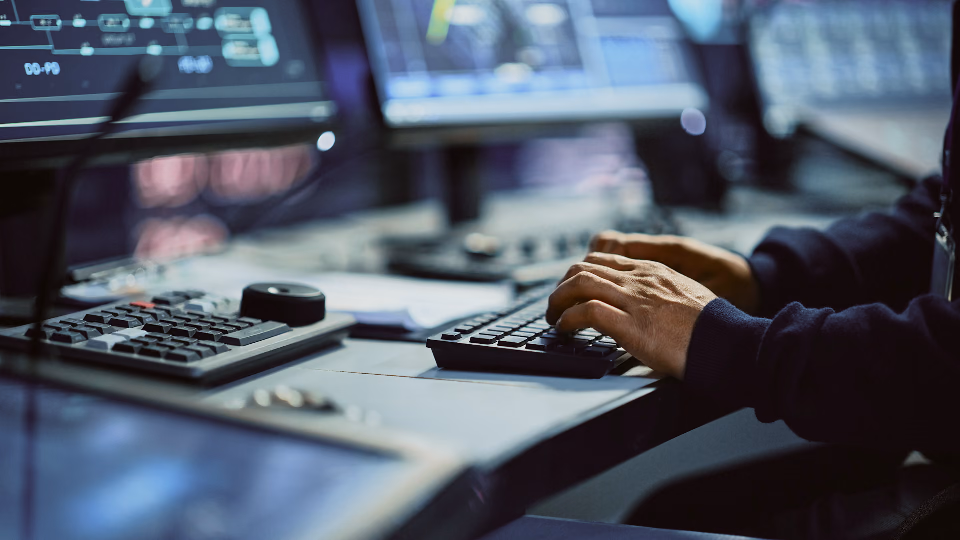Person typing on keyboard in a dimly lit room with computer screens showing data in the background.