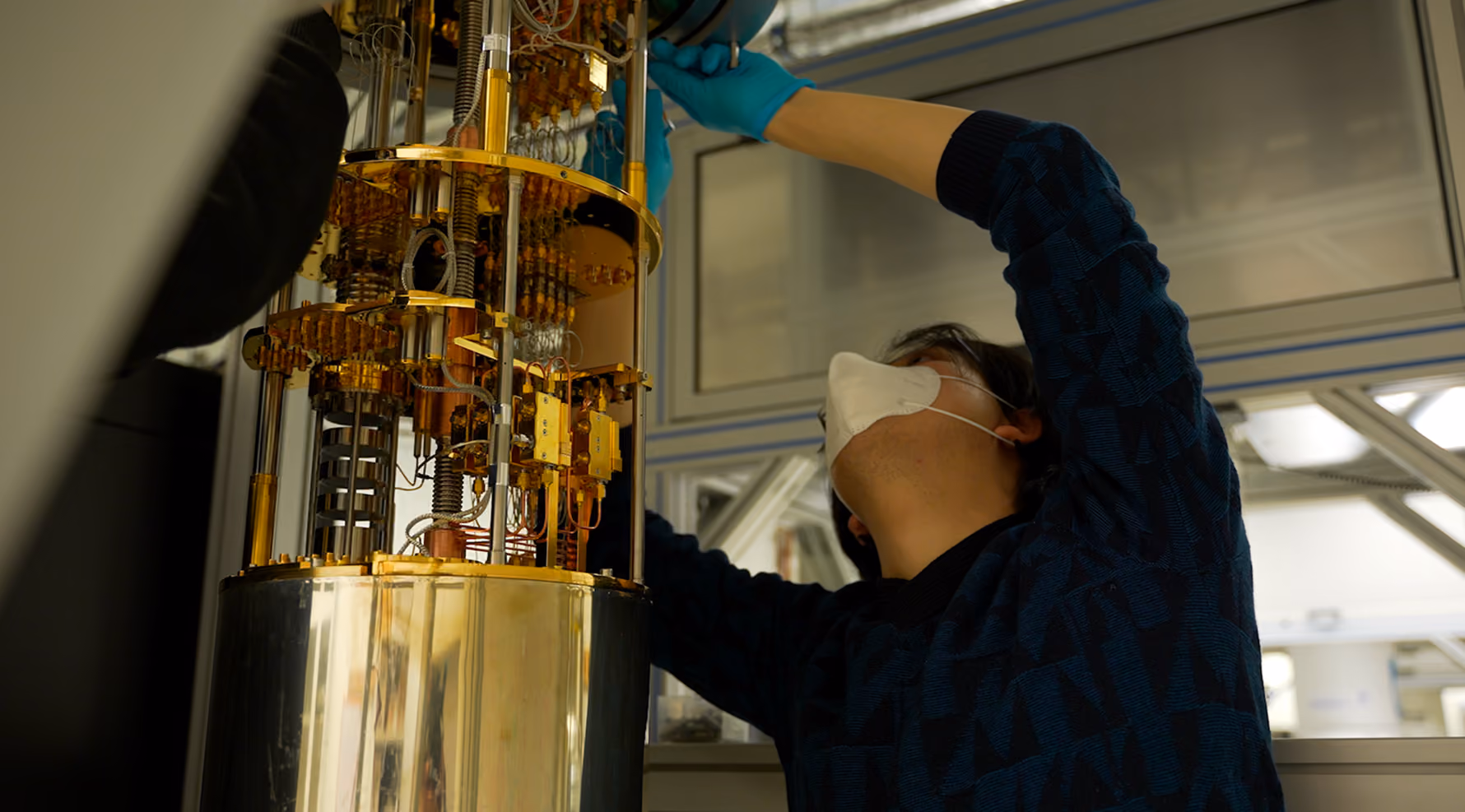An engineer in a face mask and blue gloves reaches up to adjust the gold-plated stages of a quantum computer's dilution refrigerator in a laboratory.