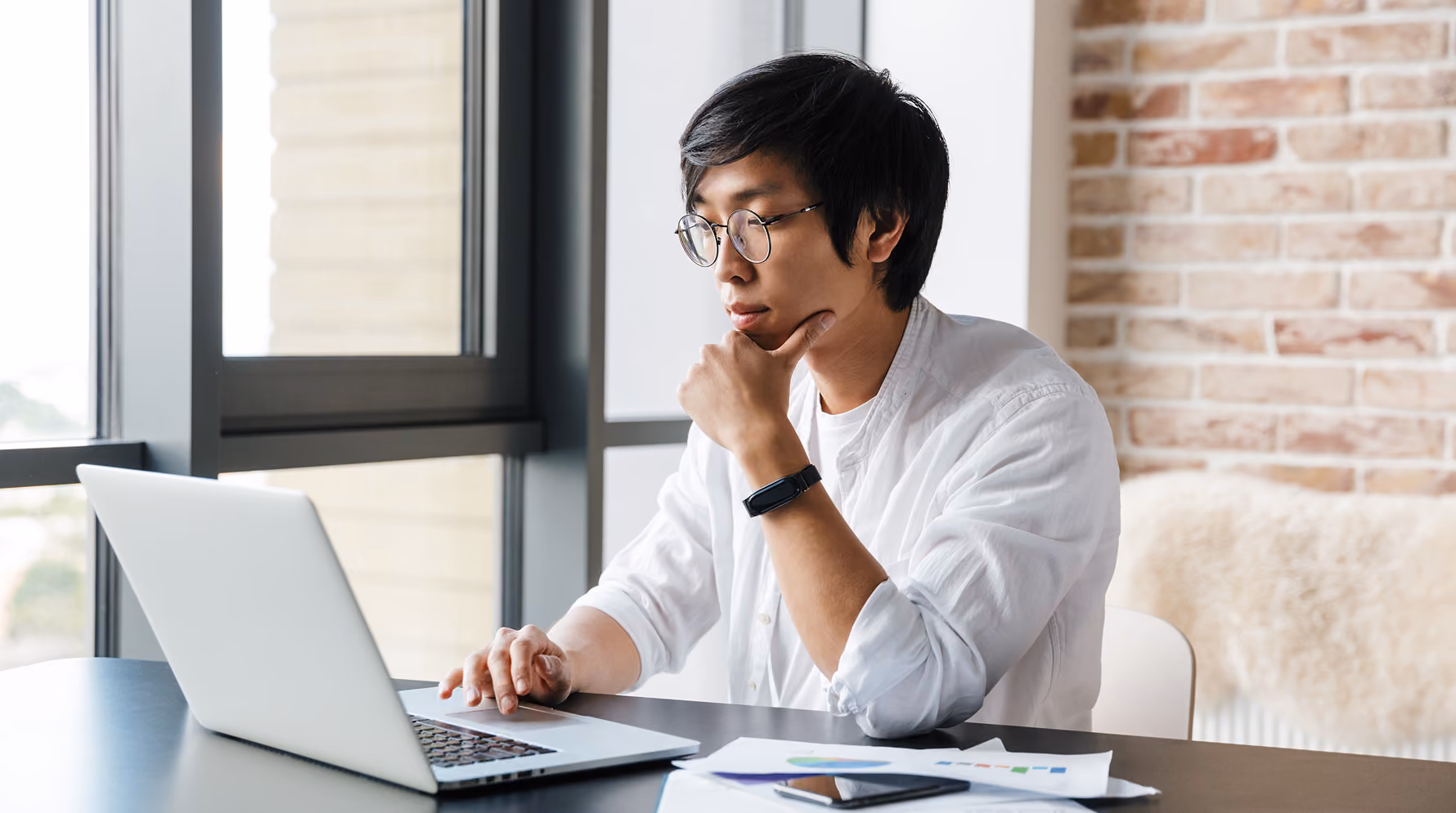 A person in glasses rests their chin on one hand while reading a laptop screen, with printed charts and a phone on the desk beside them.