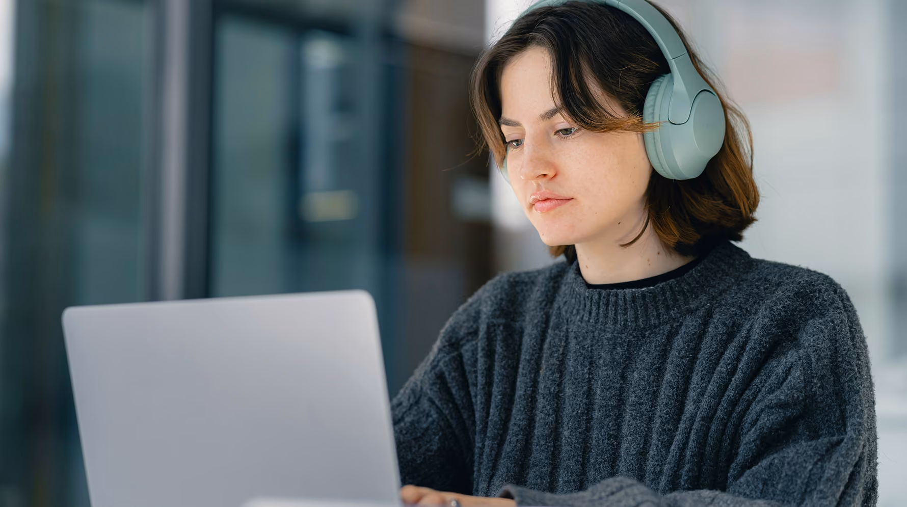 A person wearing over-ear headphones and a grey knitted jumper, concentrating on a laptop screen in a bright modern office.