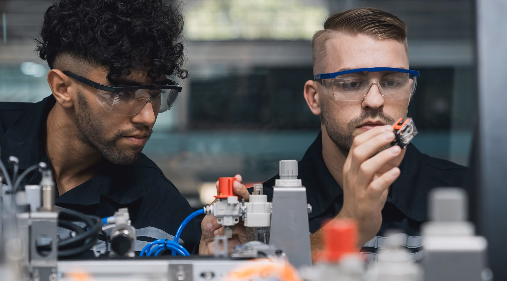 Two engineers wearing safety glasses inspect equipment on a laboratory bench, one holding a small component up to examine it while the other adjusts a valve.