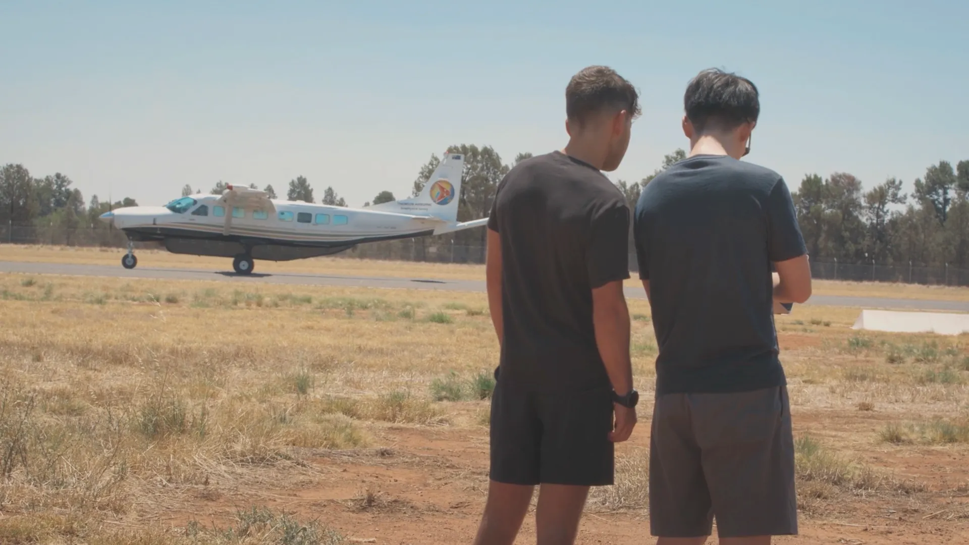 Two men standing on dry grass near a small airplane on a sunny day at an airstrip.