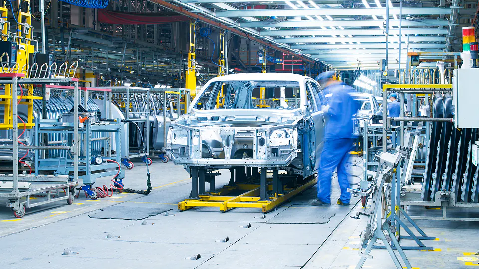 Car frames on assembly line in factory with workers in blue uniforms moving around.