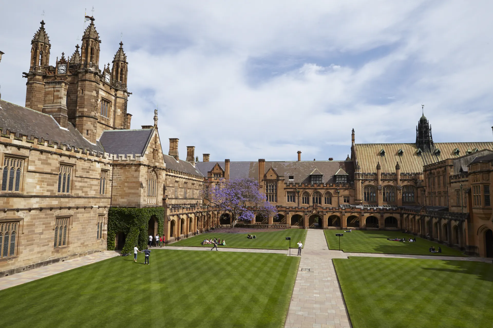University quad with green lawns, stone pathways, old Gothic-style buildings, and students sitting outdoors.