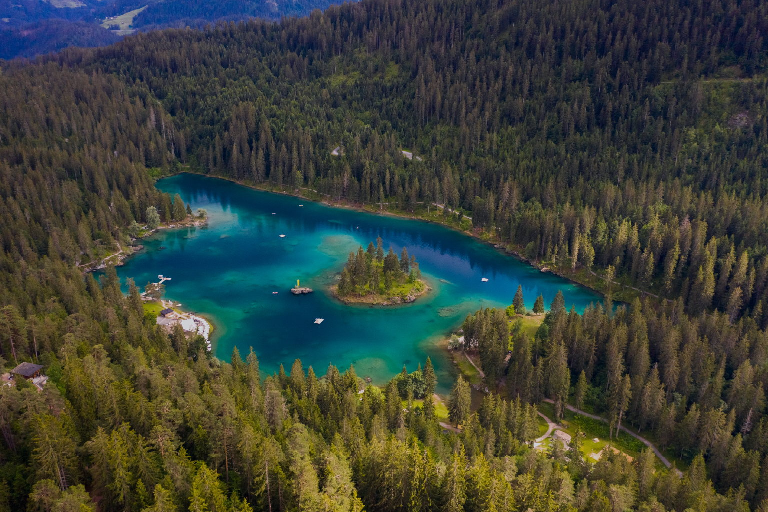 Aerial view of a small lake surrounded by dense forest with a tree-covered island in the center and several small docks or platforms on the water edges.