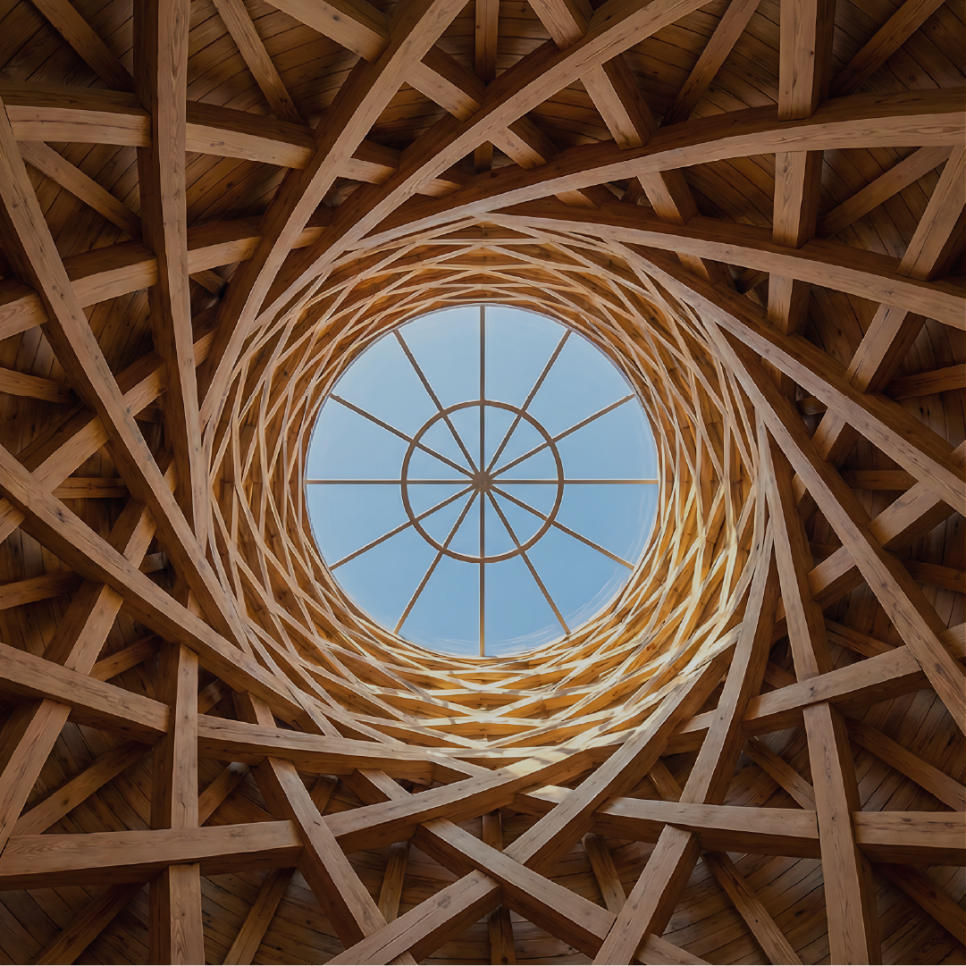 Interior view looking up through a wooden spiral lattice structure with interlocking beams forming a circular skylight at the top revealing a clear blue sky.