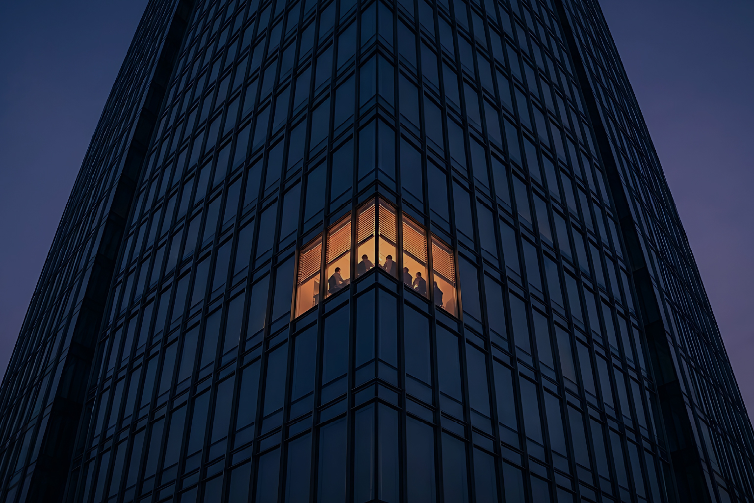 Corner of a tall glass office building at dusk with a warmly lit meeting room showing six people seated and engaged in discussion.