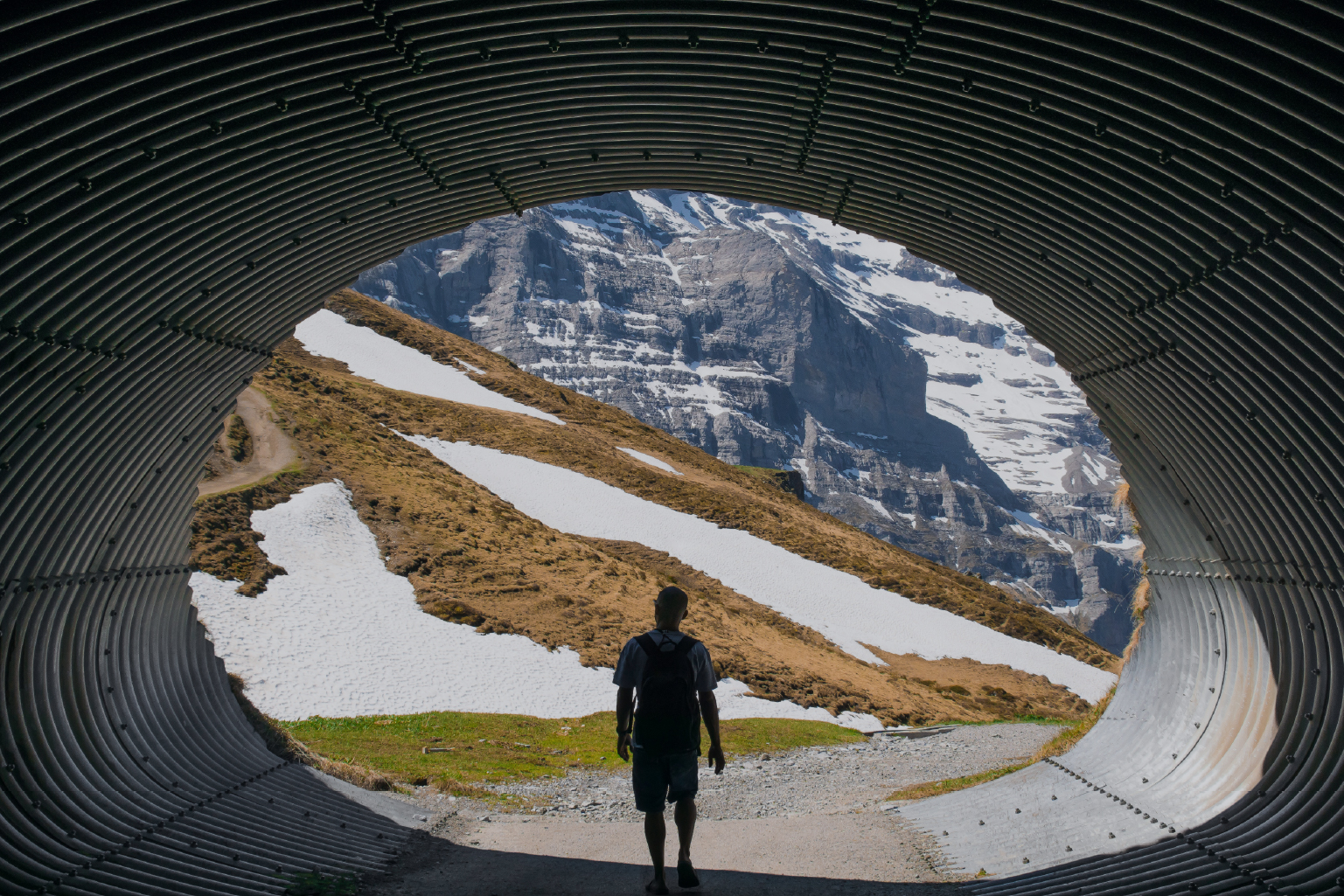 Person with a backpack walking through a corrugated metal tunnel towards a mountain landscape with patches of snow and grass under a clear sky.