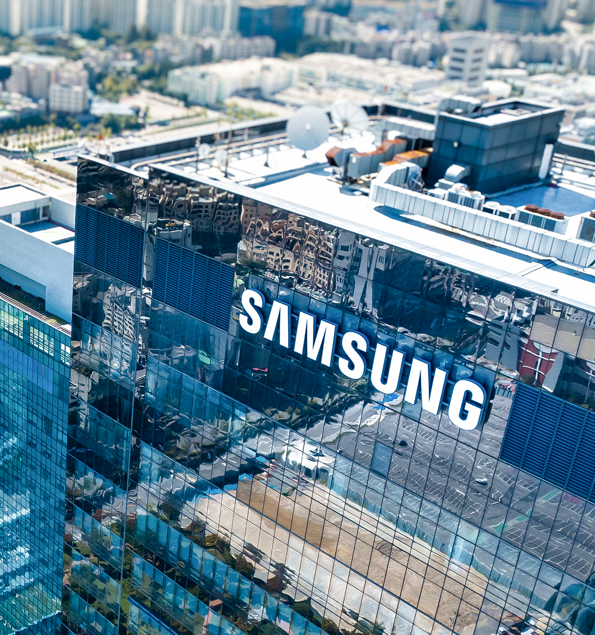 High-angle view of a modern glass office building with the Samsung logo prominently displayed on the facade, reflecting surrounding city buildings and streets.