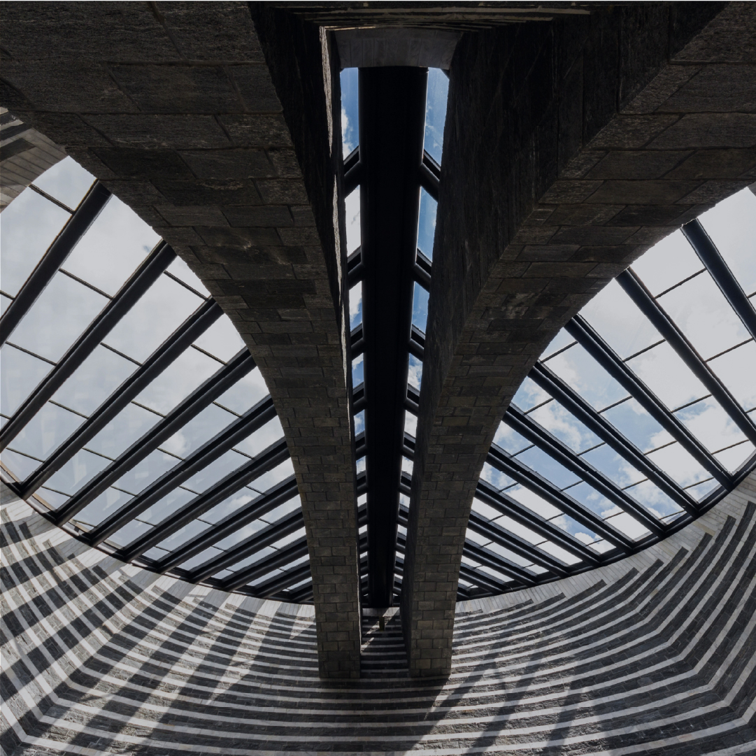 Interior view of a modern architectural structure featuring two large, curved stone arches supporting a glass roof with visible sky and clouds above, creating striped shadow patterns on the curved stone walls below.