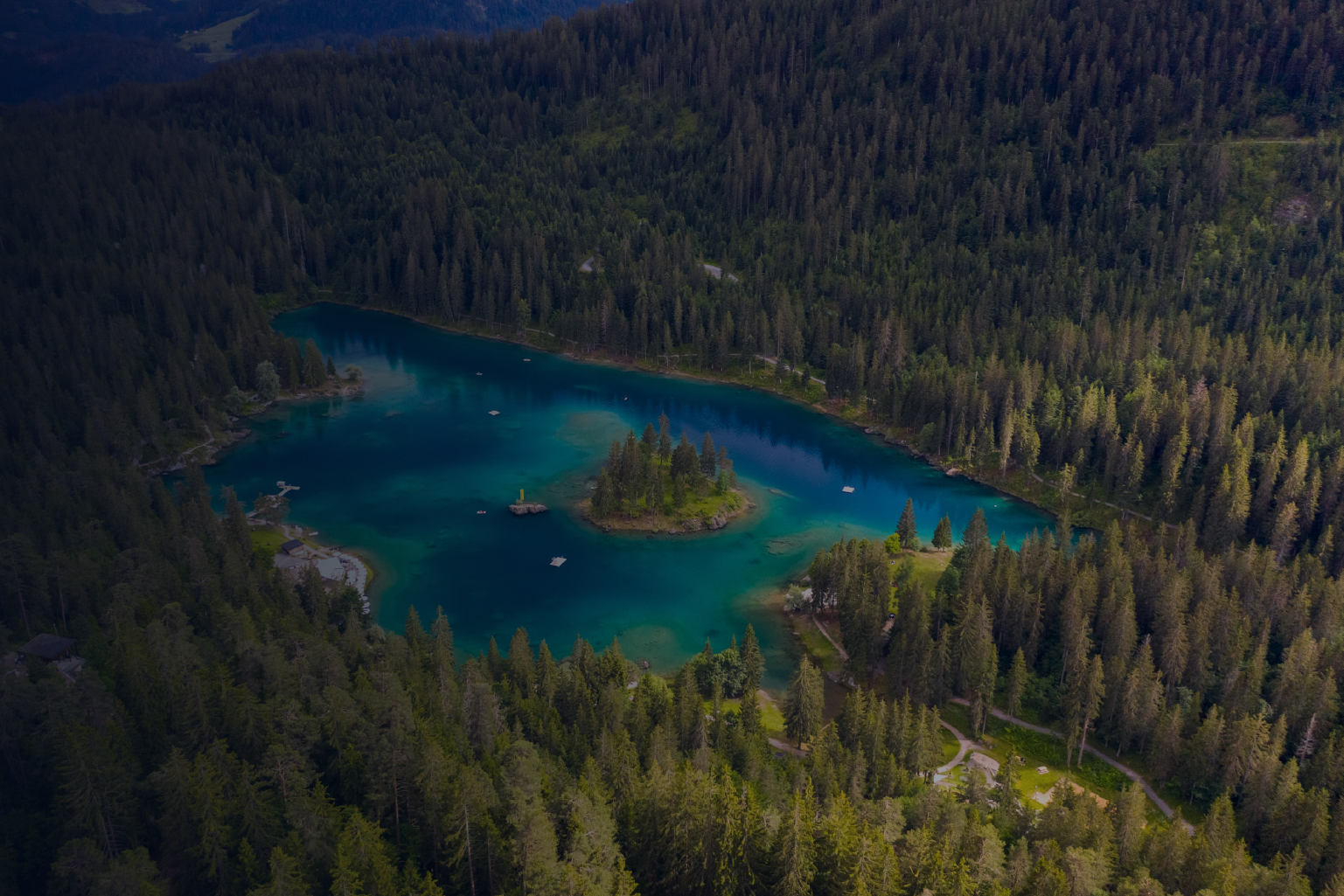 Aerial view of a small lake surrounded by dense forest with a tree-covered island in the center and several small docks or platforms on the water edges.