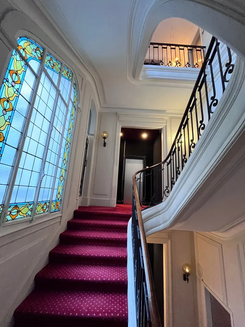 Palier d’immeuble rénové avec grand vitrail et escalier à tapis bordeaux, Paris 16e