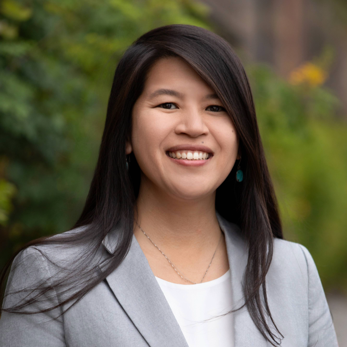Primer plano de una mujer con collar de plata, blusa blanca y traje gris claro. Sonríe al aire libre.
