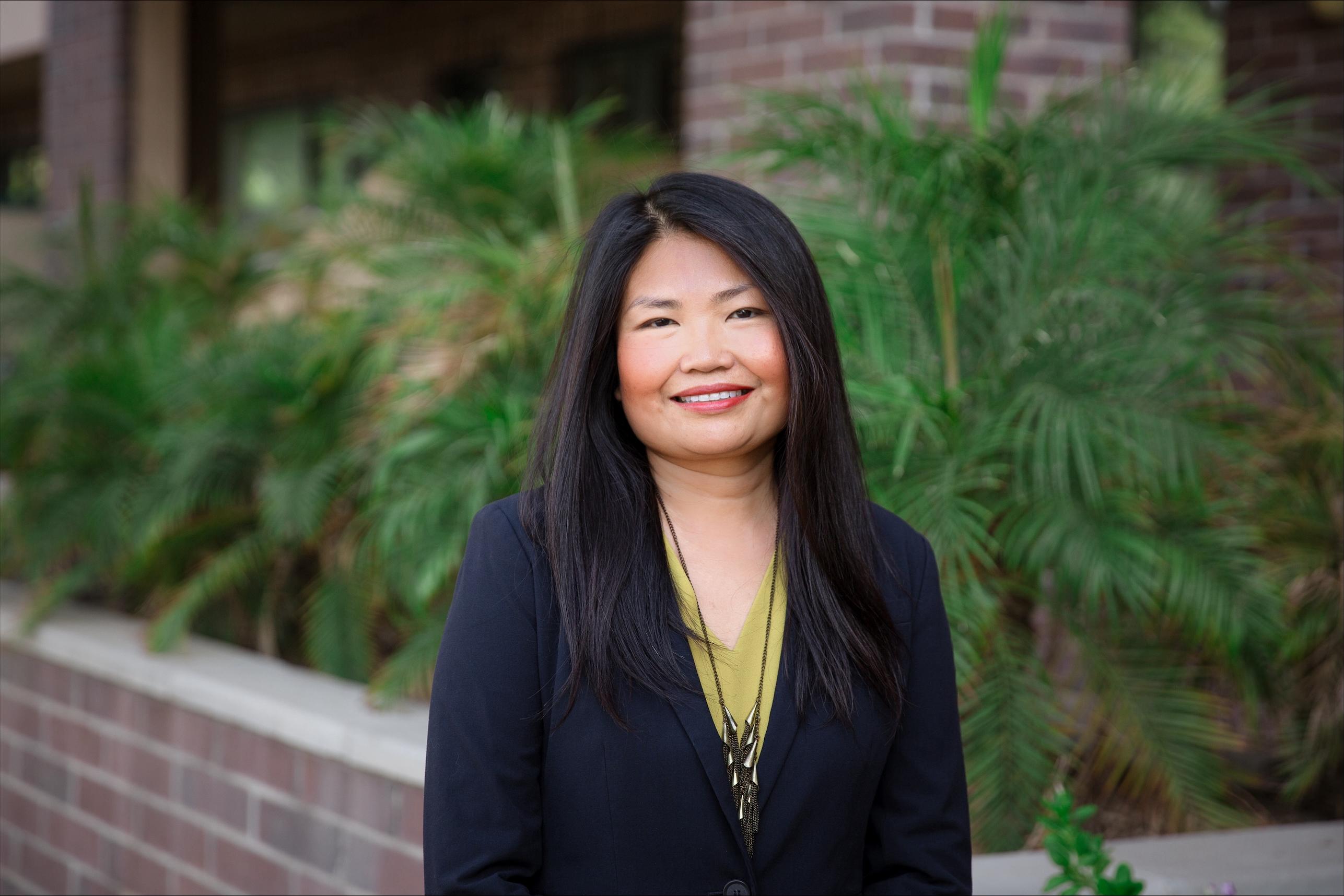 Primer plano de una mujer sonriente con blusa verde y traje negro. Está de pie frente a una valla de ladrillos, con las palmas en la espalda.