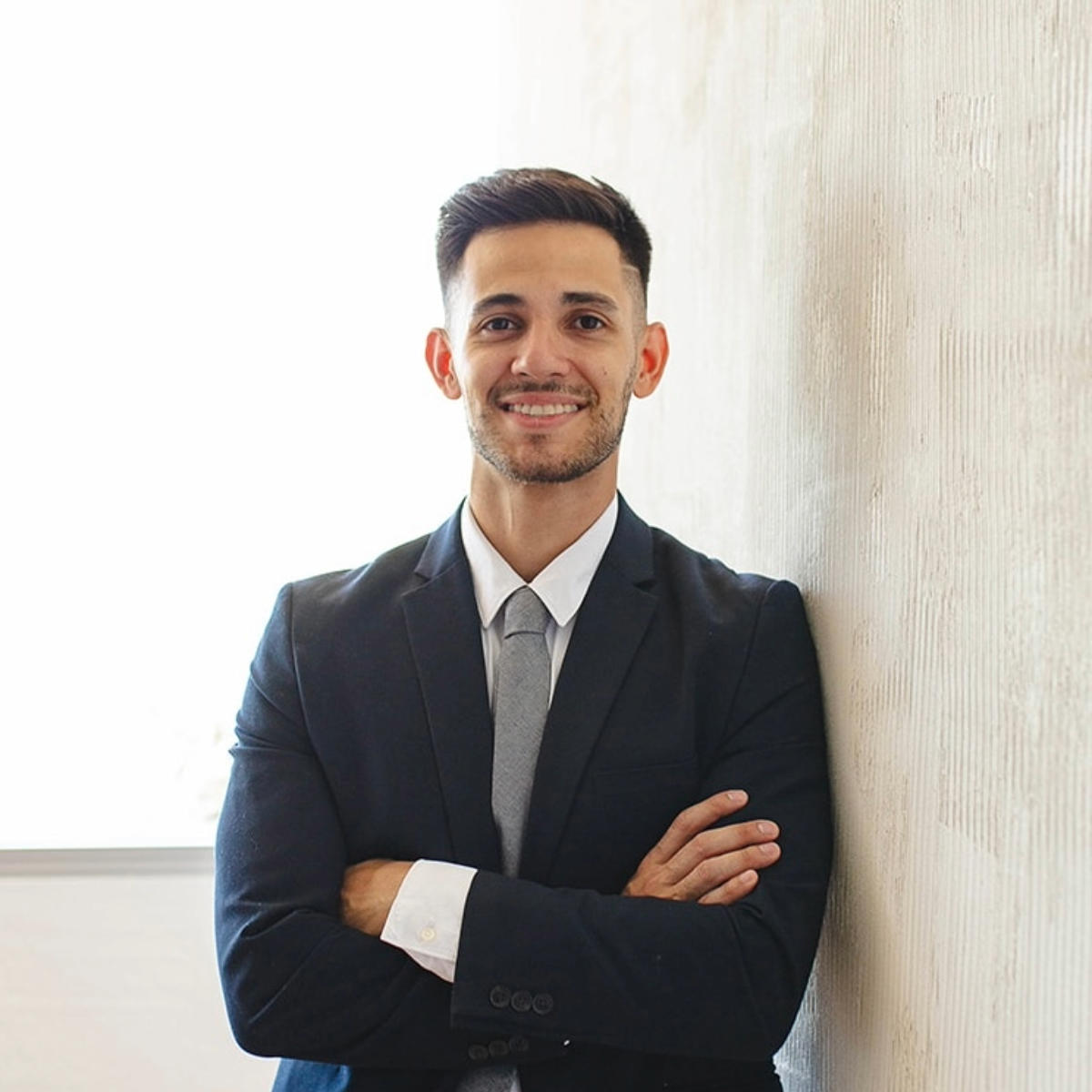 Un hombre vestido de traje, de pie en una habitación blanca con pisos de madera, se apoya contra una pared y sonríe.