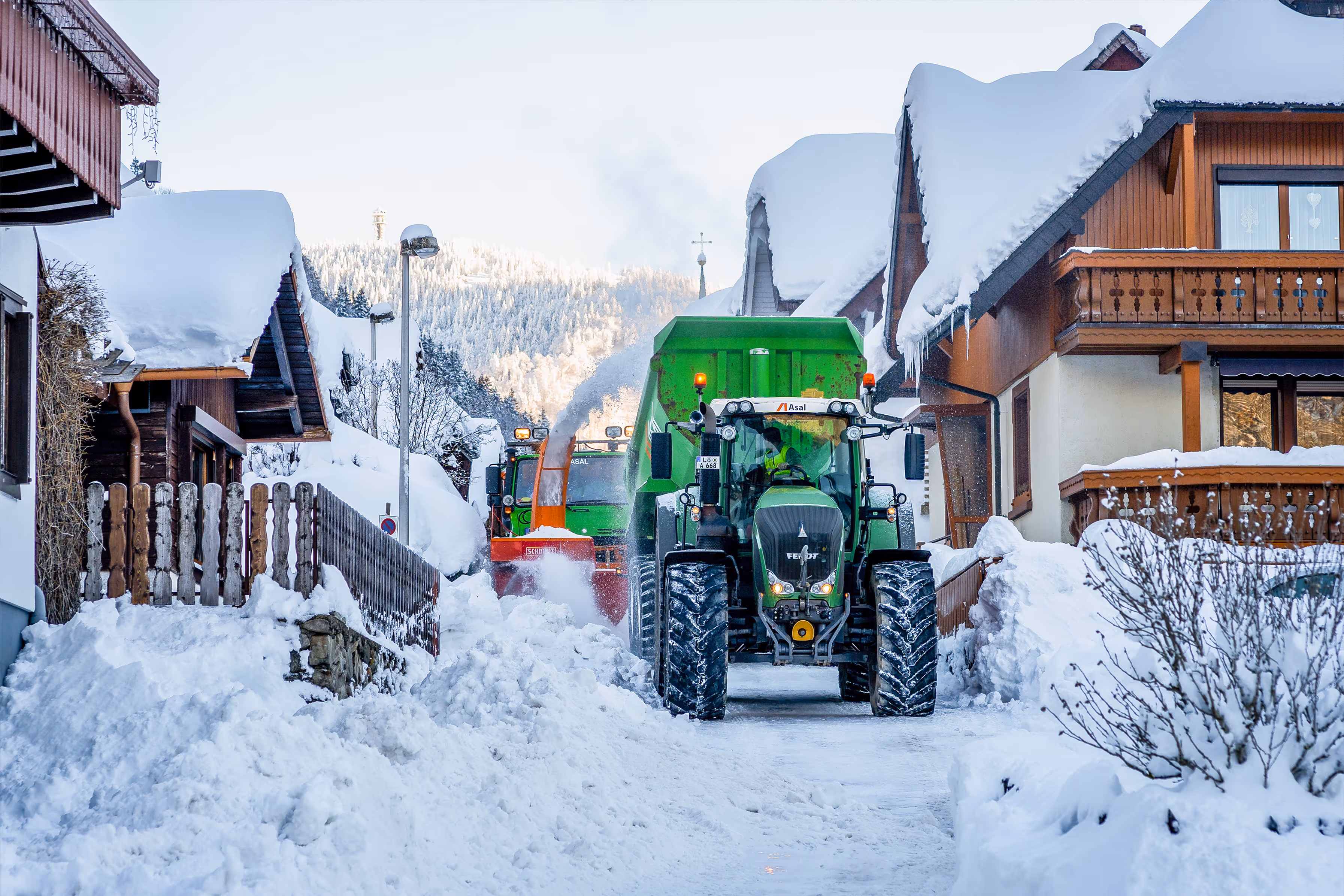 Traktor mit Schneefräse räumt Straße zwischen verschneiten Häusern, Schnee wird in Anhänger geschleudert.