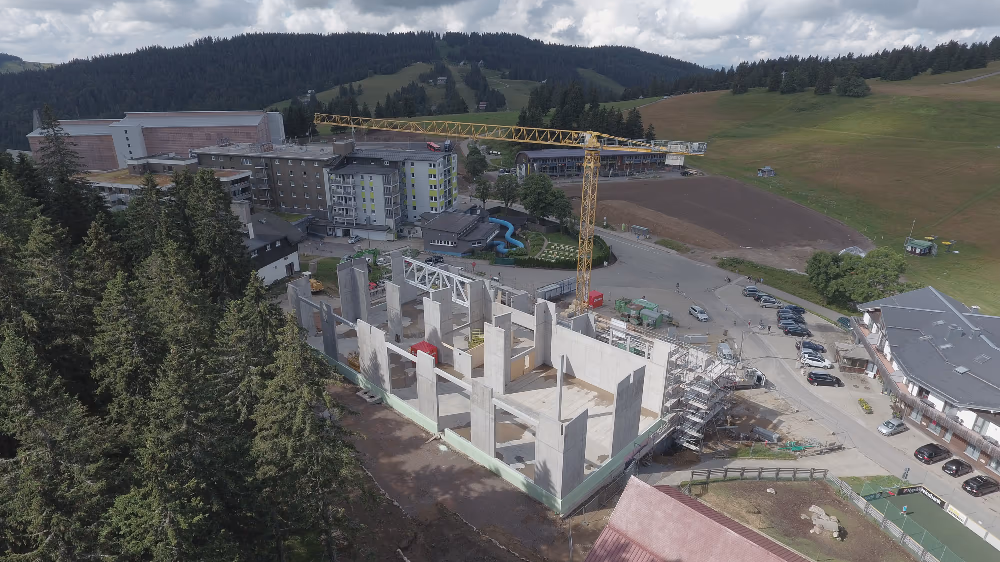 Neubau der "Fundorena" auf dem Feldberg. Baustelle eines Rohbaus im Gebirge mit gelbem Baukran, umgeben von Hotels, Straßen und Parkplätzen.