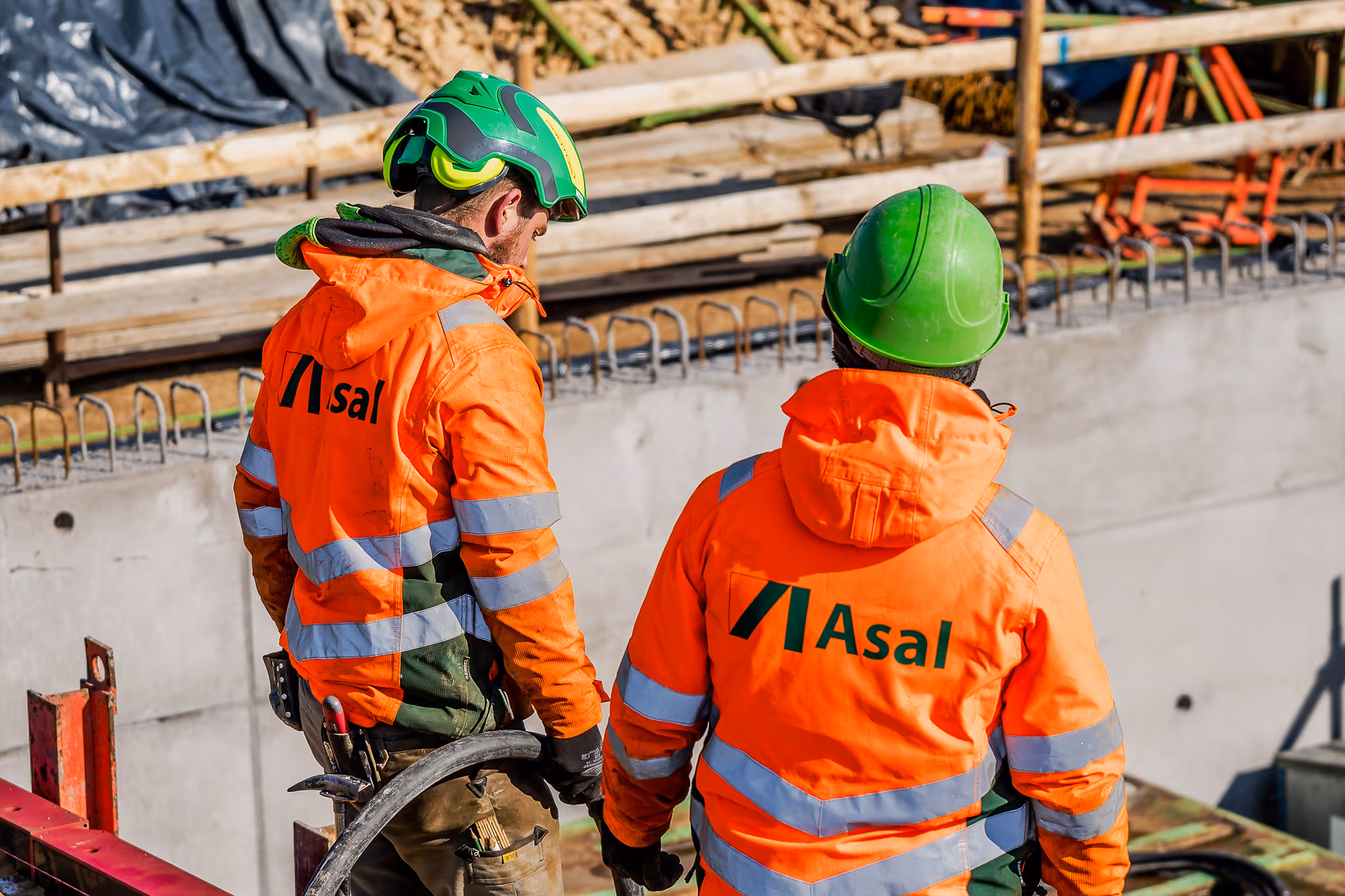 Zwei Bauarbeiter mit Asal-Jacken und grünen Helmen arbeiten an einer Baustelle.