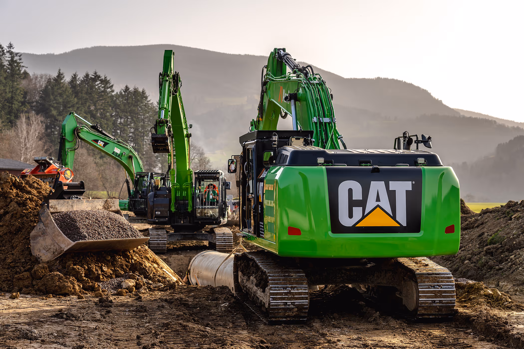 Mehrere grüne Bagger von Asal und CAT bei Tiefbauarbeiten auf einer Baustelle im Tal, im Vordergrund Erdaushub und Rohrleitungen