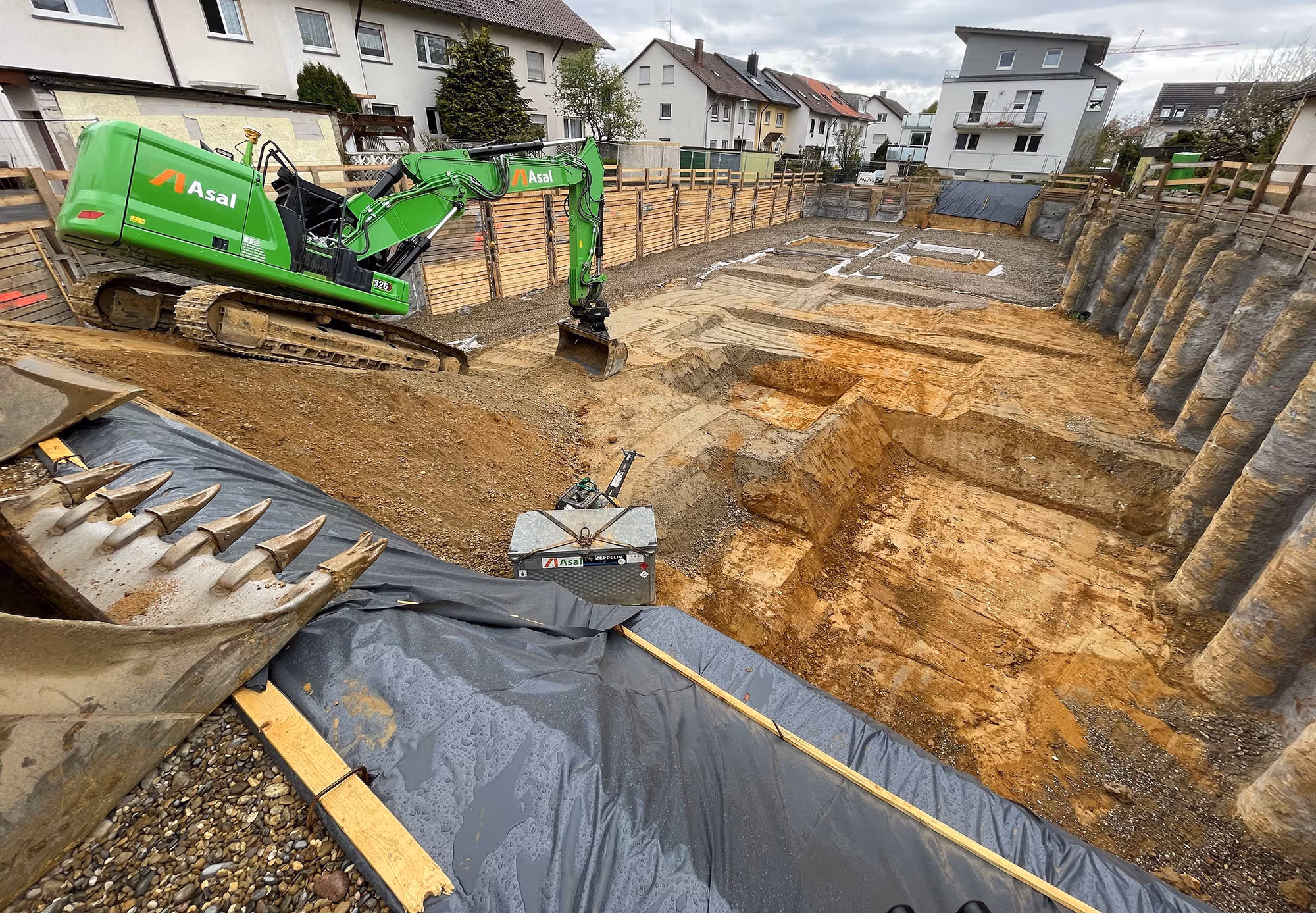 Grüner Asal-Bagger belädt einen Traktor mit Erdreich auf einer innerstädtischen Baustelle, dahinter Wohnhäuser