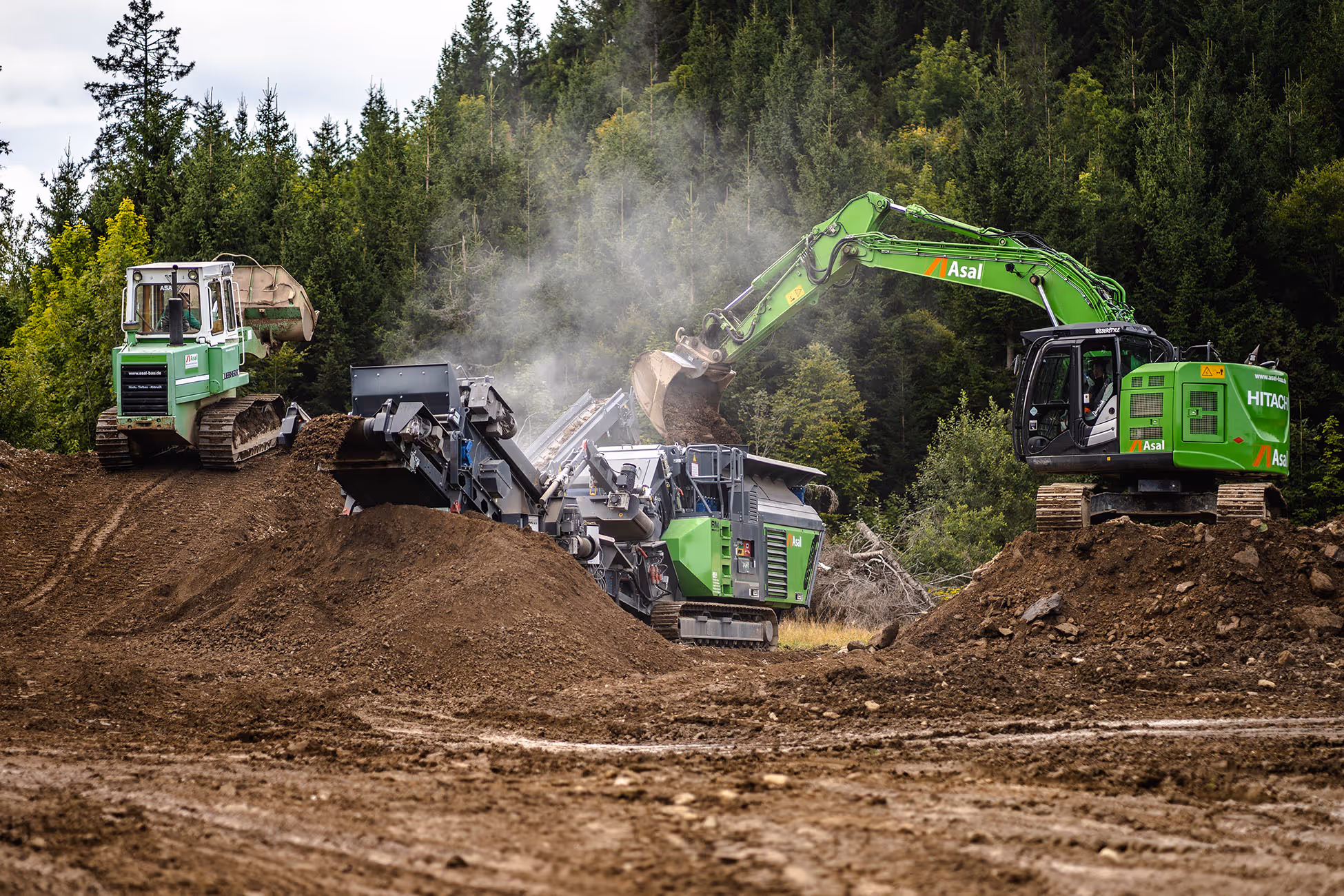 Bagger mit Greifer lädt Erde und Gestein in mobile Brechanlage, im Hintergrund ein Raupenlader auf einem Erdwall