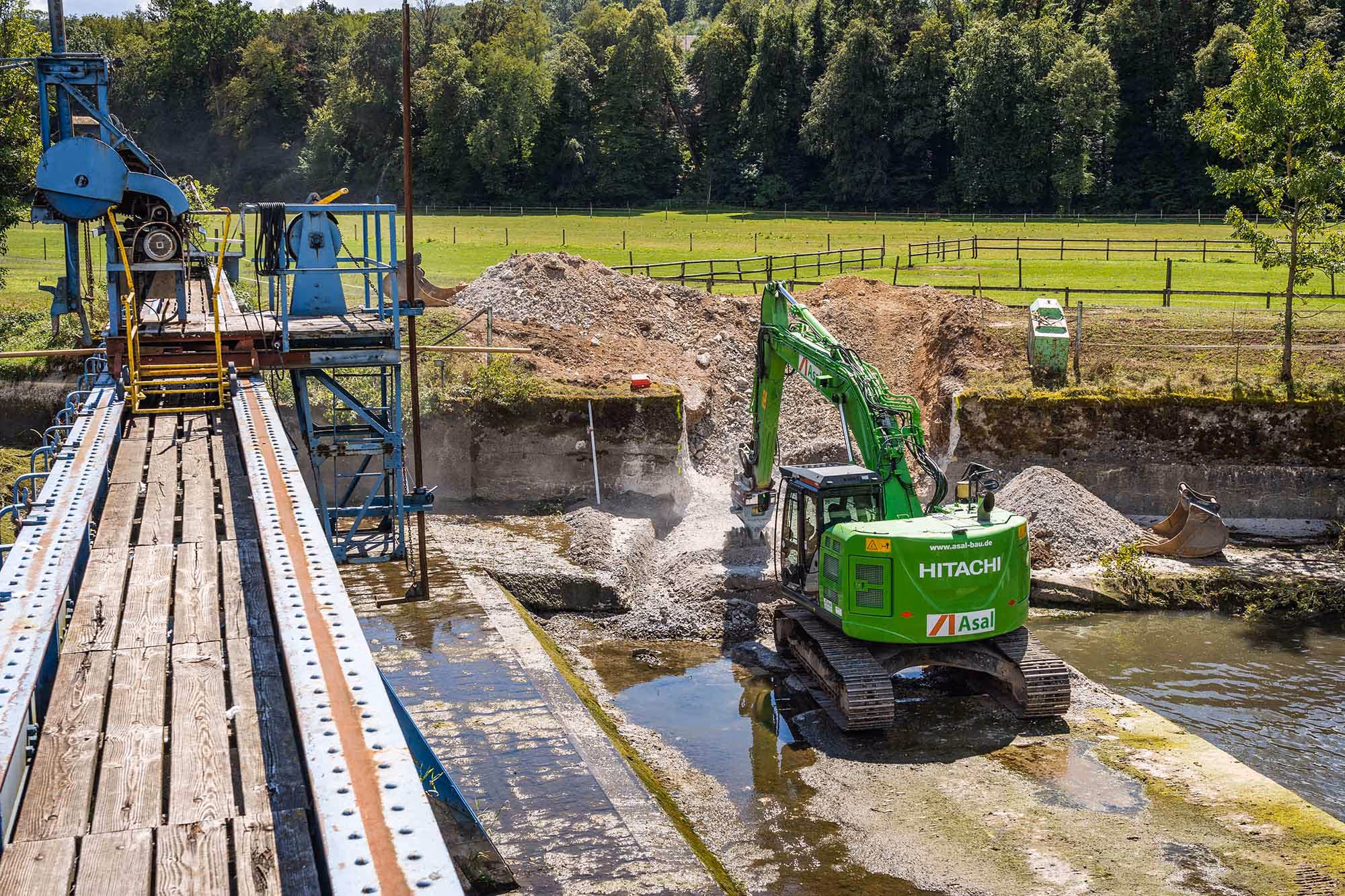 Bagger führt Rückbauarbeiten an Betonbauwerk im Flussbereich aus, Baustelle an einem Gewässer.