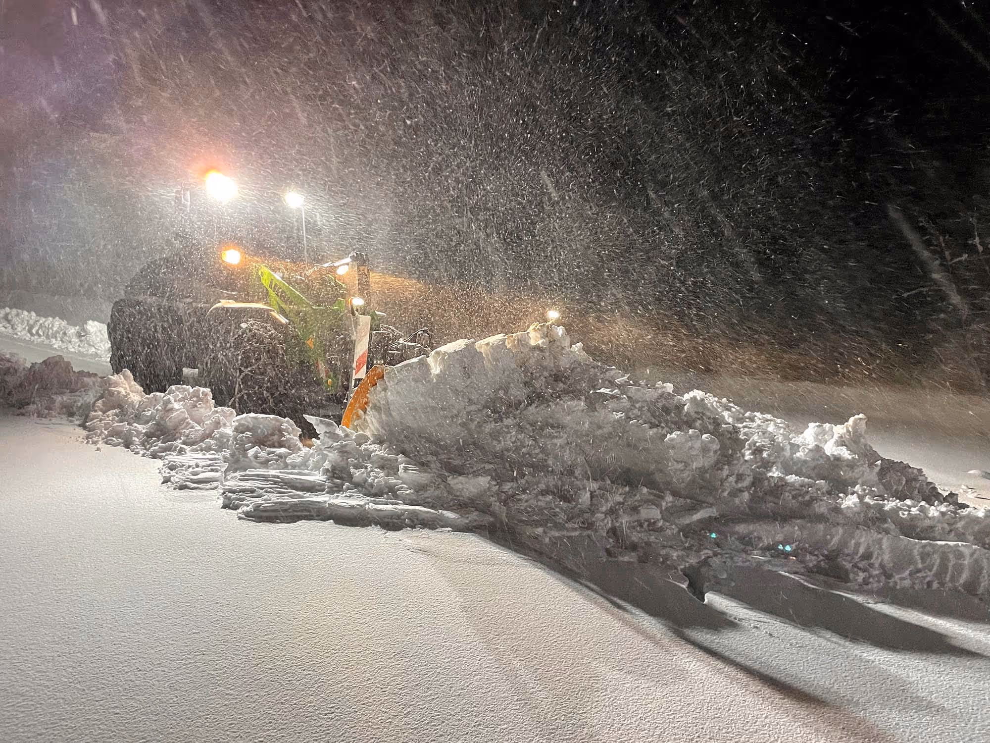 Radlader mit Schneeschild räumt in der Nacht bei starkem Schneefall eine Straße frei.