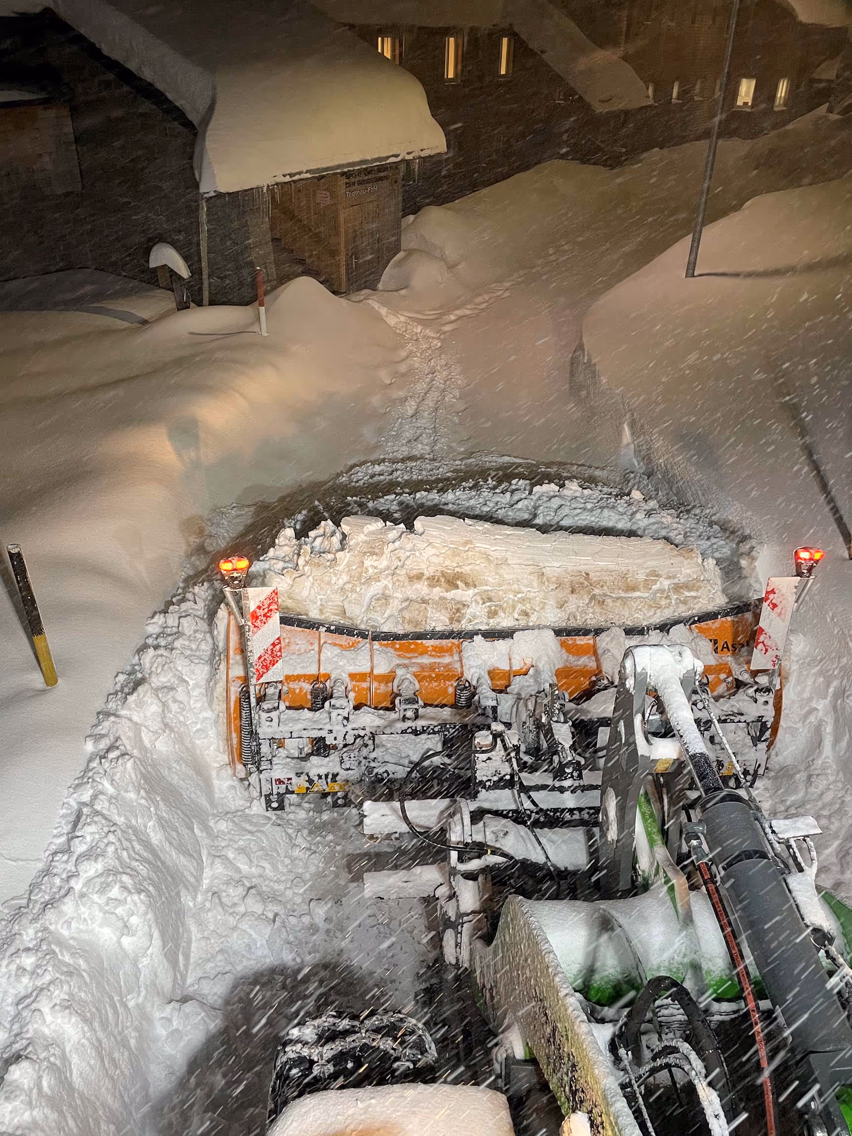 Schneepflug in nächtlichem Einsatz räumt engen Weg zwischen hohen Schneemauern.